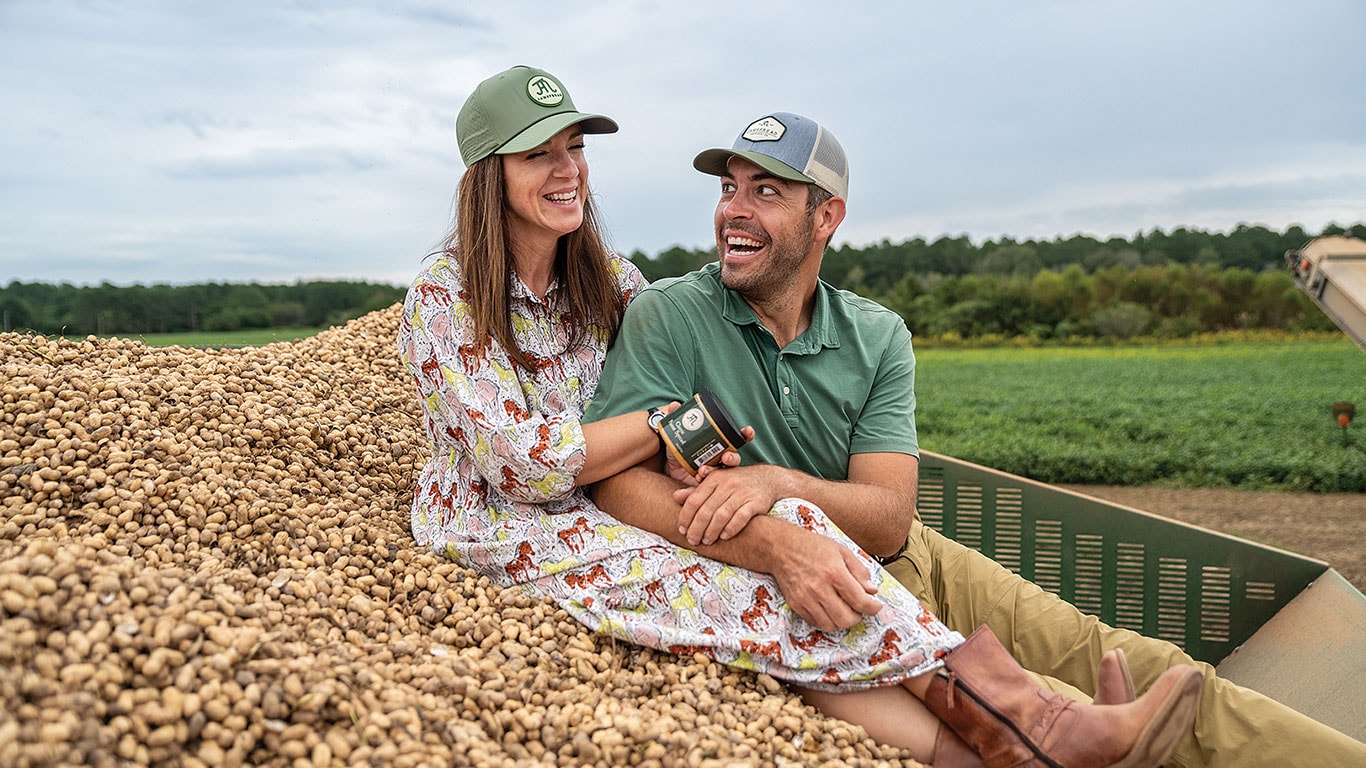 Two people sitting on a large pile of peanuts in a farm field with green crops in the background.