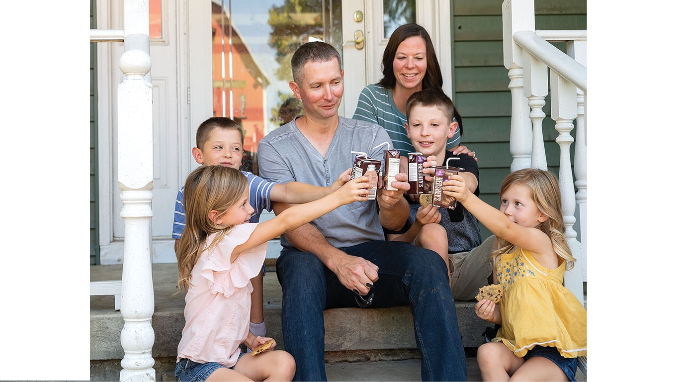 A group of children and adults sit on porch steps, joyfully raising chocolate milk cartons.