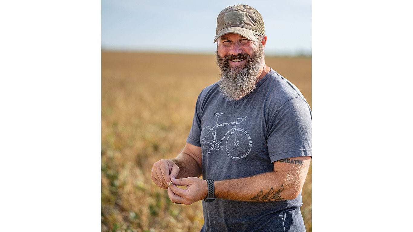Person in a gray T-shirt and cap standing in a field, holding and inspecting a plant pod.
