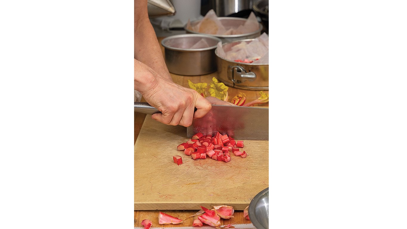 A hand cuts bright pink rhubarb on a wooden cutting board.