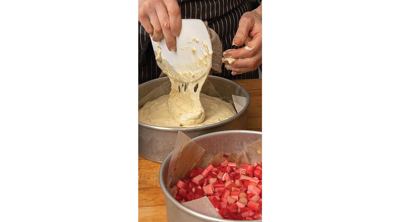 A person pours cake batter into a round baking pan, a bowl with chopped rhubarb is nearby.