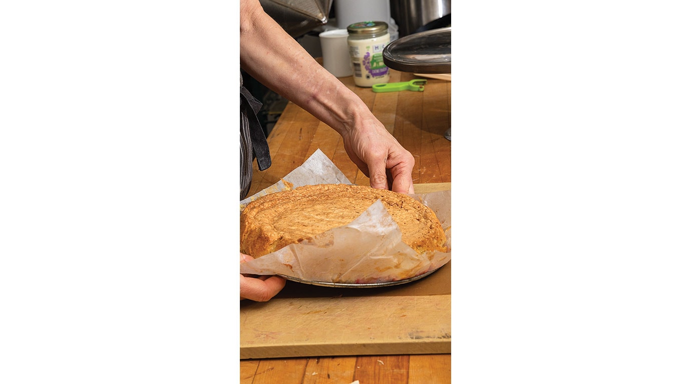 A person is carefully removing a freshly baked cake from a round pan lined with parchment paper.