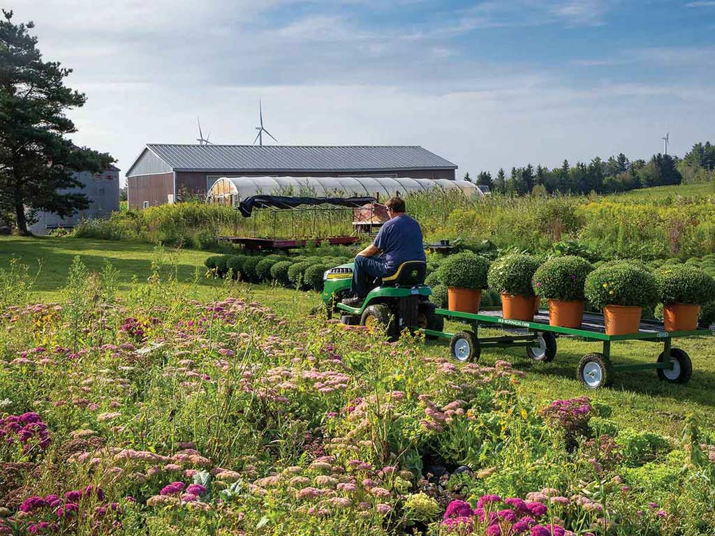 Person on a lawn tractor hauling a row of potted plants through a blooming garden