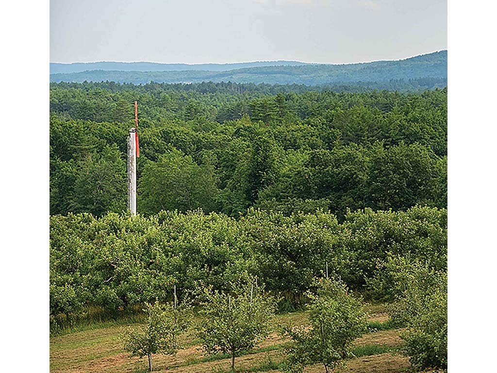 A tall post with colorful markings stands amidst lush green orchards and rolling hills under a cloudy sky.