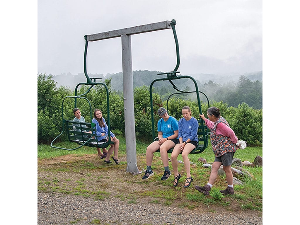 Two children sit on a green ski lift chair, while a woman stands nearby.