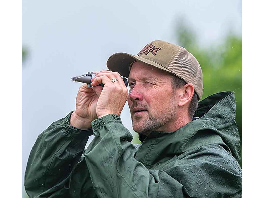A person in a green rain jacket and a tan cap holds a small device, in a rainy outdoor setting.