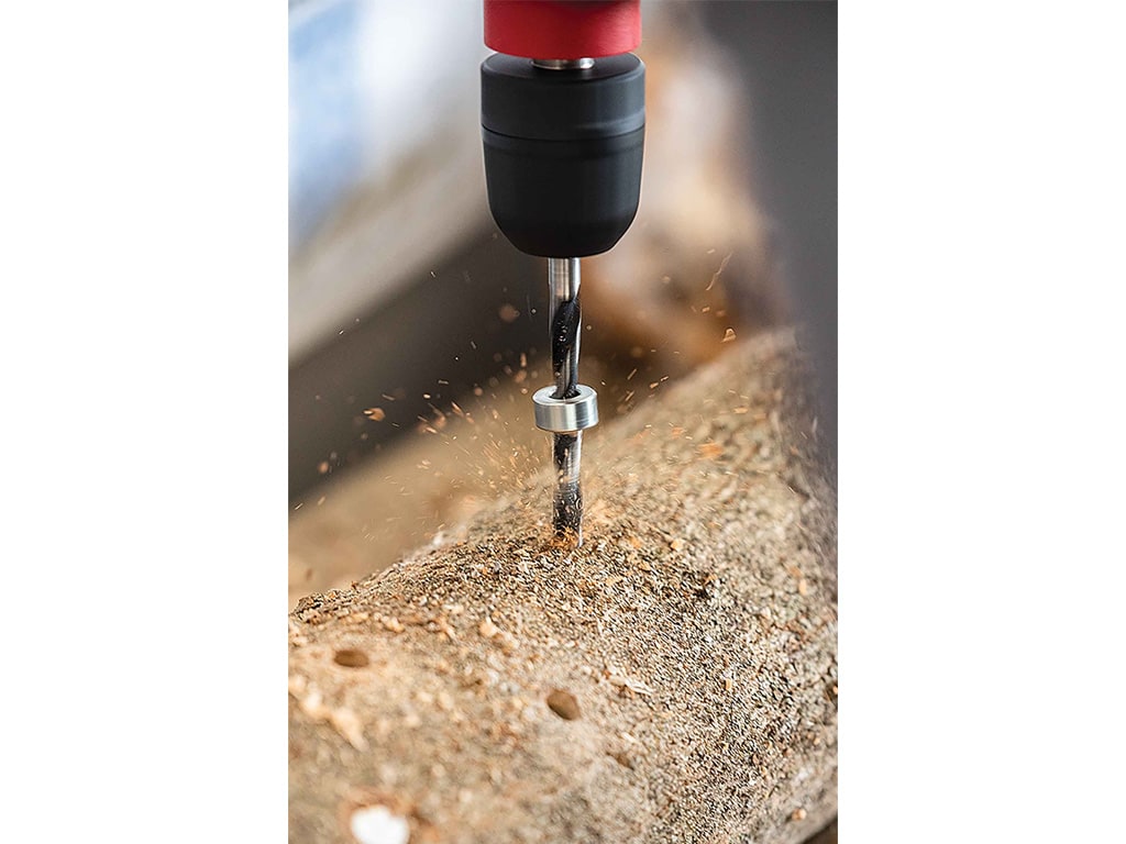 A close-up of a drill bit penetrating wood, with wood shavings flying off.