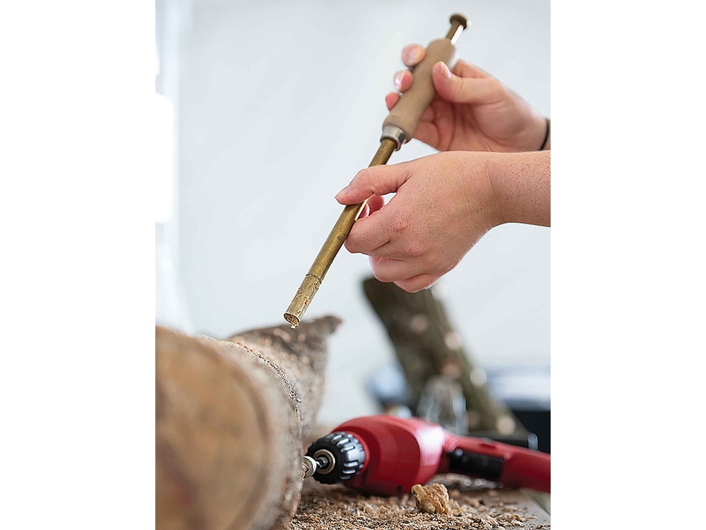 A person holds a brass tool above a wooden log, with a red drill nearby.