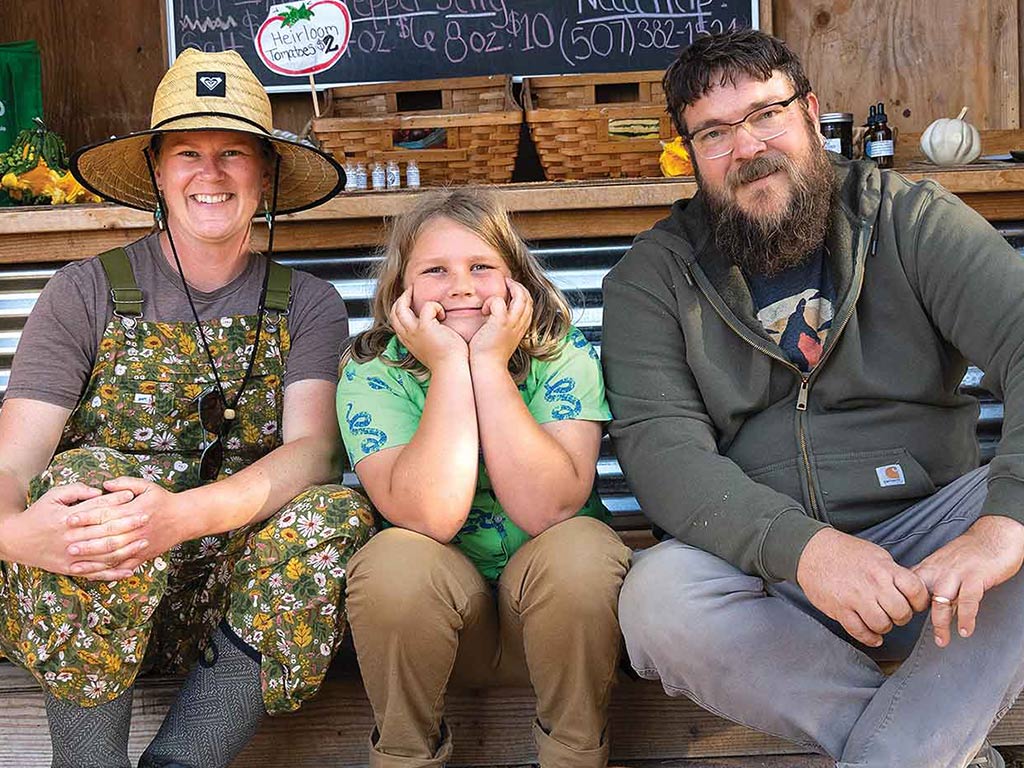 A person in a straw hat and overalls sits beside a child in a green shirt, with another adult in a gray hoodie on the other side.