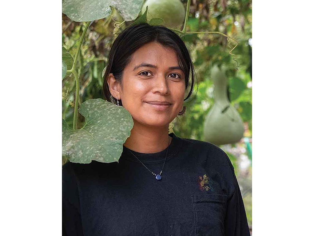 A young person stands amidst green foliage, partially obscured by large leaves and hanging gourds.