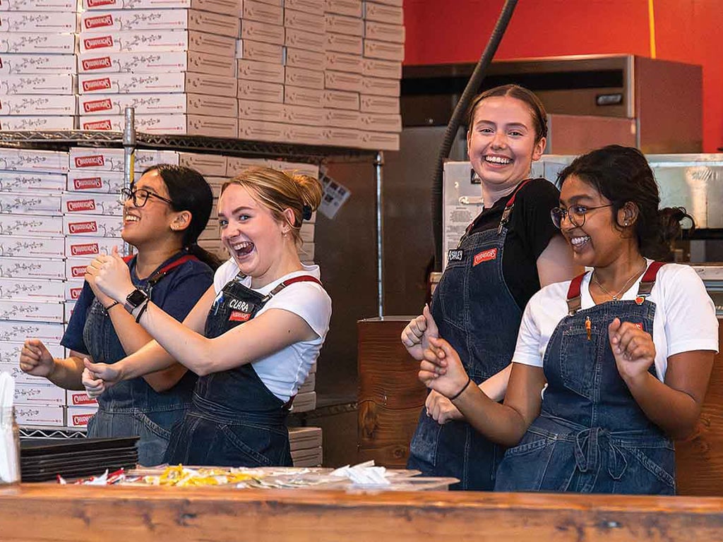 A group of restaurant employees wearing denim overalls dances together behind a counter filled with pizza boxes and condiments.
