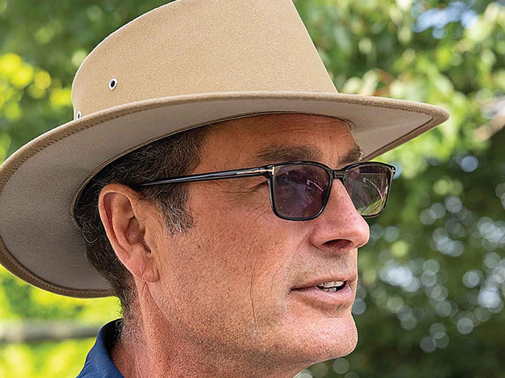 A man with glasses and a wide-brimmed beige hat stands outdoors, surrounded by soft green foliage and dappled sunlight.