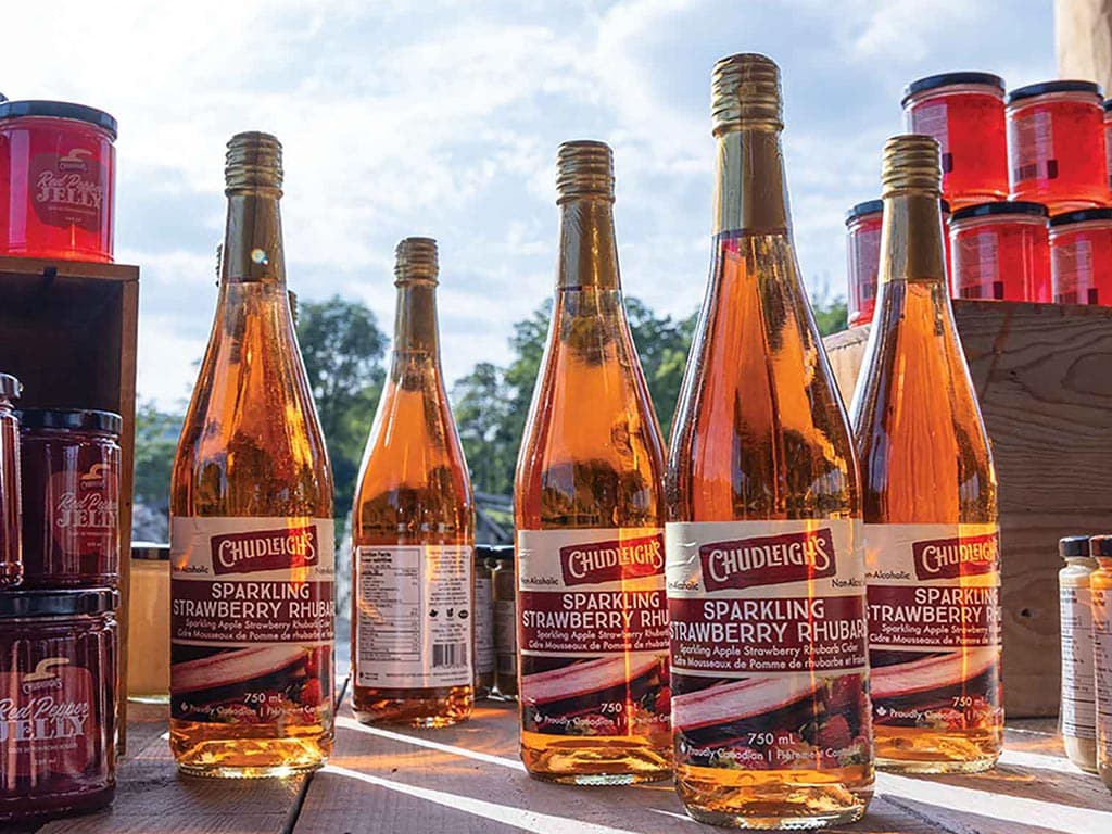 Five bottles of Chudleigh's Sparkling Strawberry Rhubarb drink are displayed next to jars of jelly under a cloudy sky.