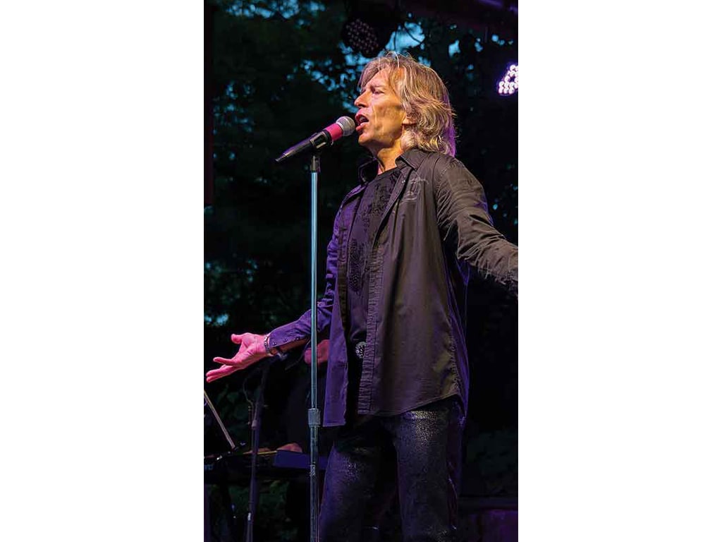 A performer in a black shirt and shiny pants passionately gestures while singing at an outdoor concert.