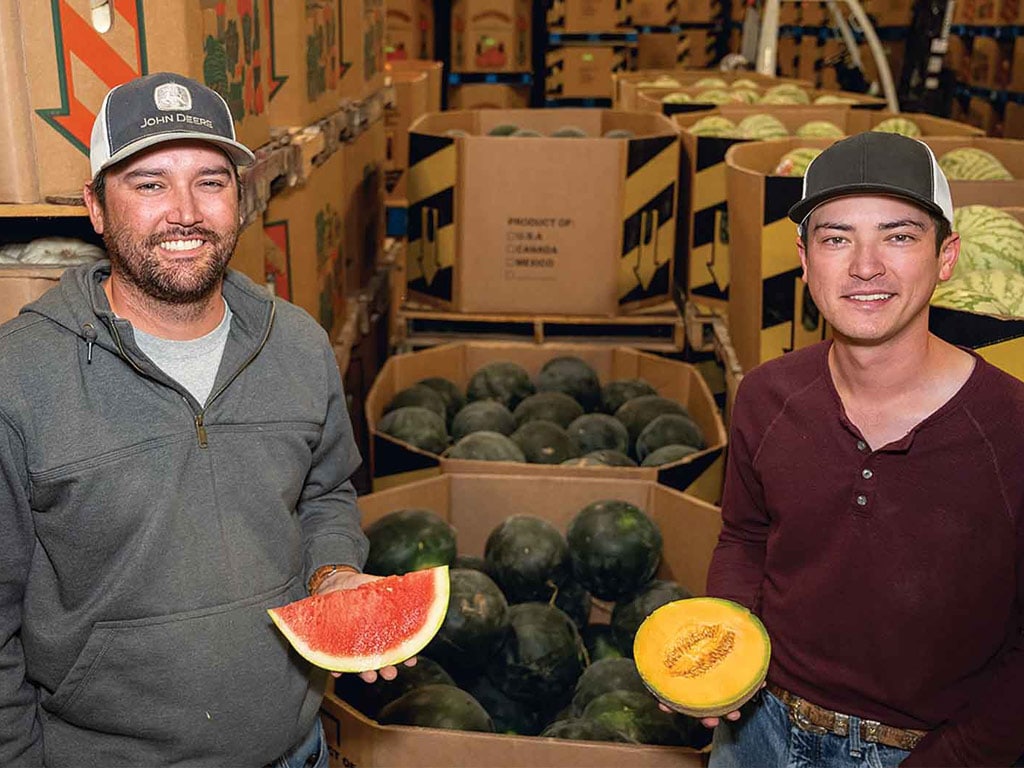 Two people standing in a produce packing area, one holding a large slice of watermelon and one holding half of a cantaloupe