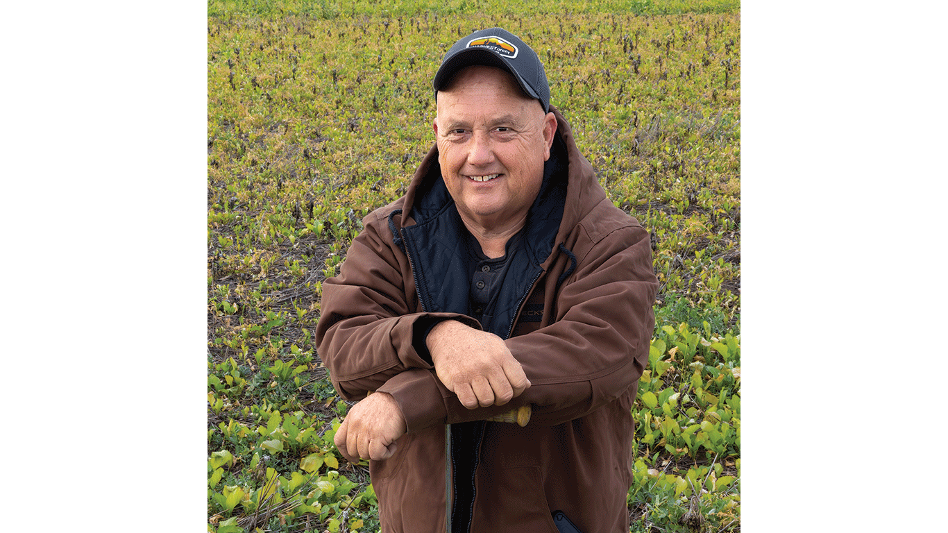 Person wearing a jacket and cap standing in a green farm field, arms crossed.