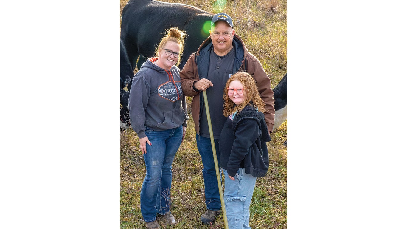 Three people standing in a grassy field with cattle behind them; one person holds a long measuring stick.