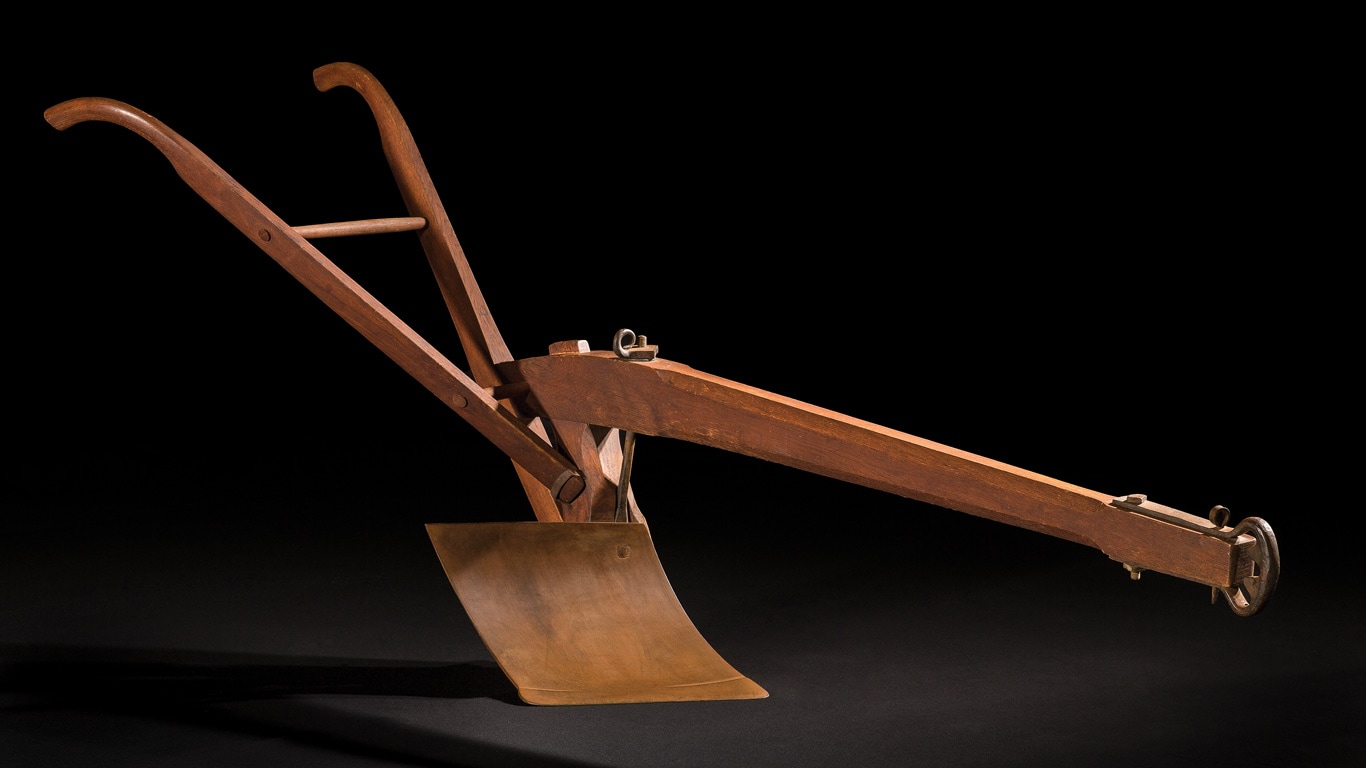 Vintage wooden agricultural plow with angled blade and handles, photographed against a black background.