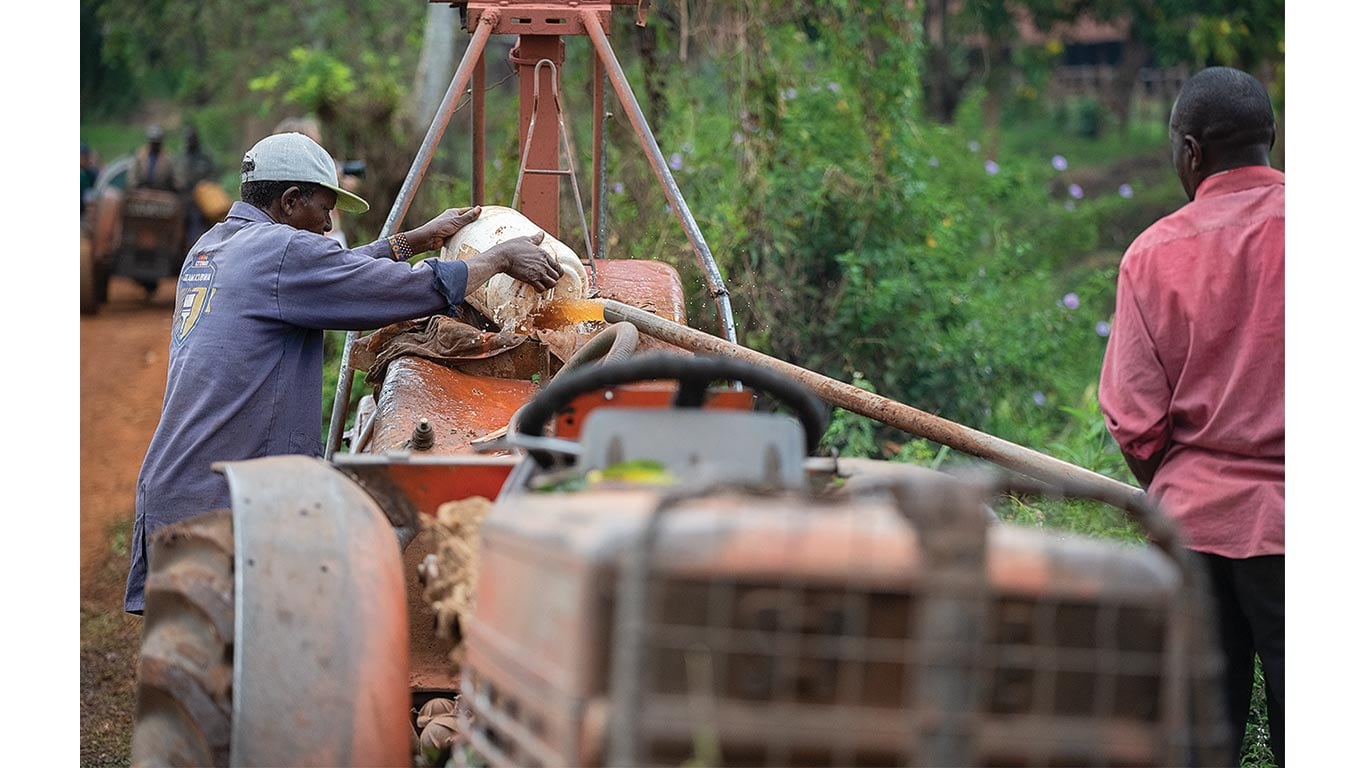 Farmer pours rinse water into a sprayer tank while another person stands nearby in a farm field.