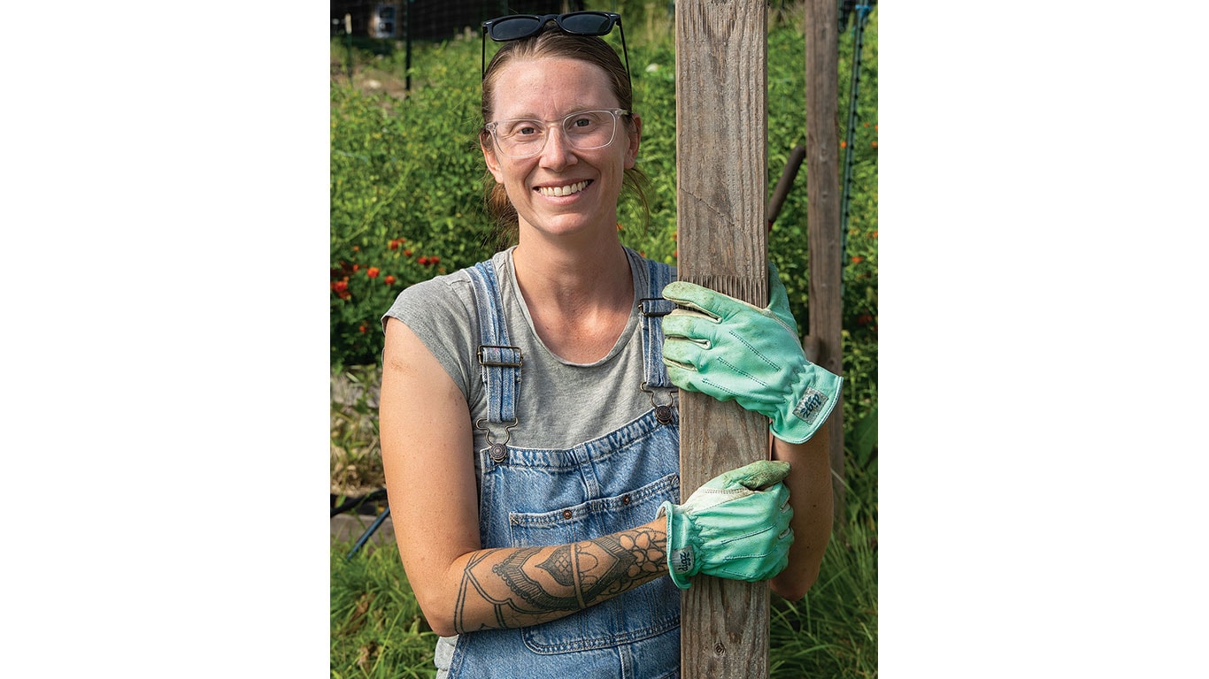 Person wearing gloves and overalls smiles while holding a wooden post in a garden.