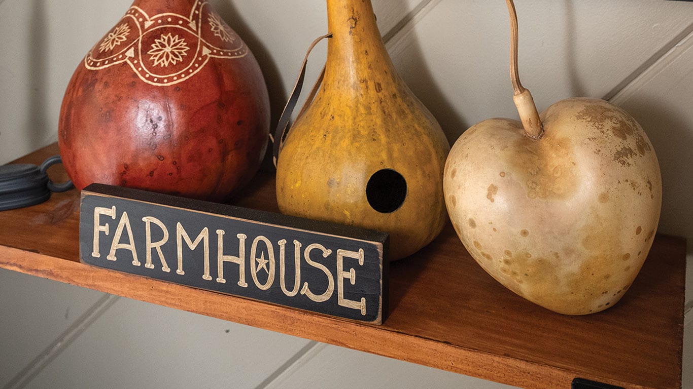 Decorative gourds displayed on a shelf with a "Farmhouse" sign.