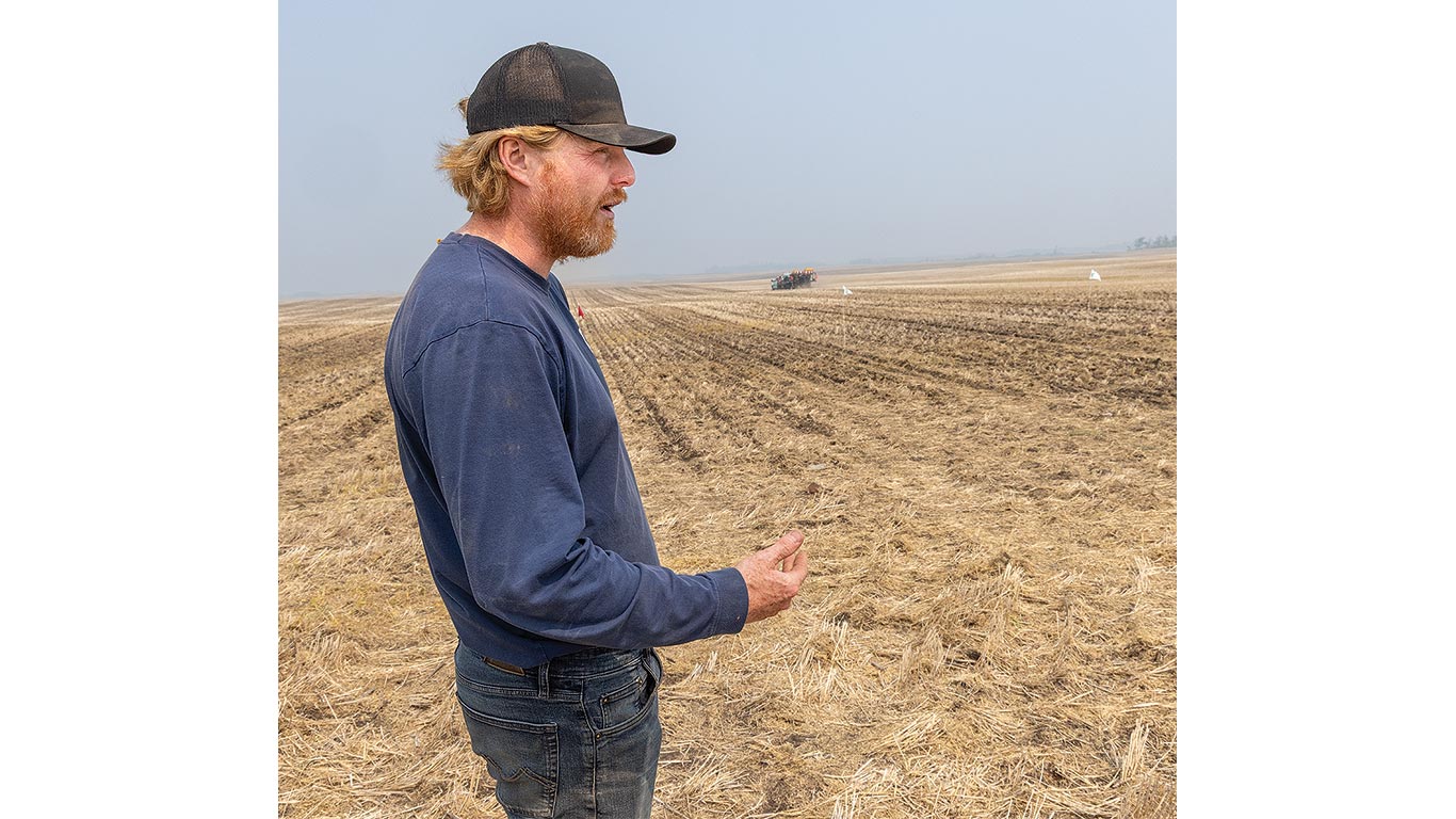 A farmer stands in a harvested field, examining soil as equipment works in the distance.