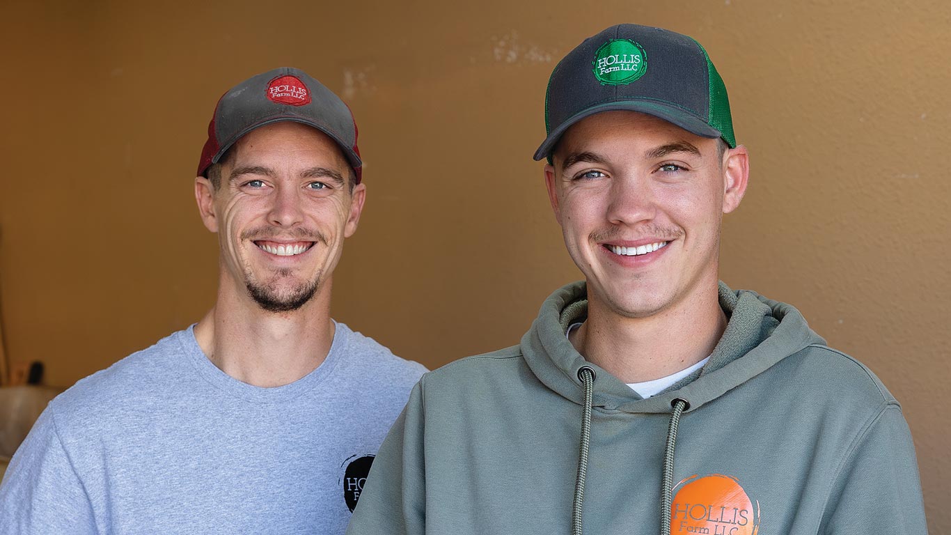 Two young men wearing farm hats smile at the camera.