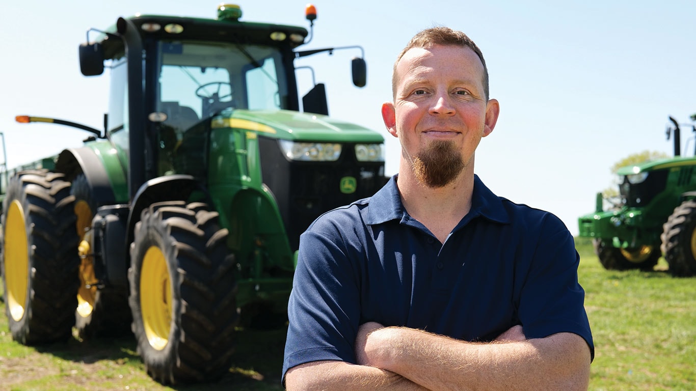 Person stands with arms crossed in a grassy field, with two large green tractors parked behind.