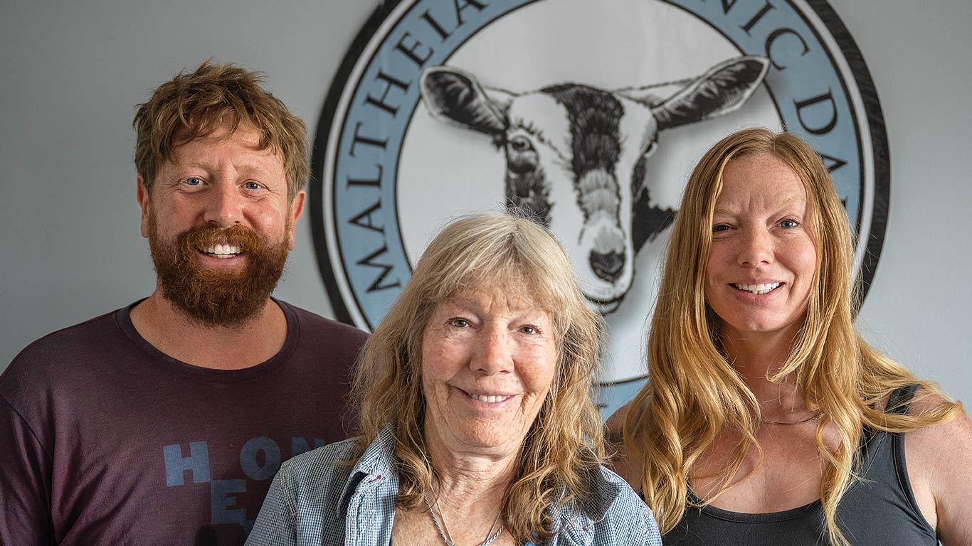 Three people stand smiling in front of a livestock logo at a dairy facility.