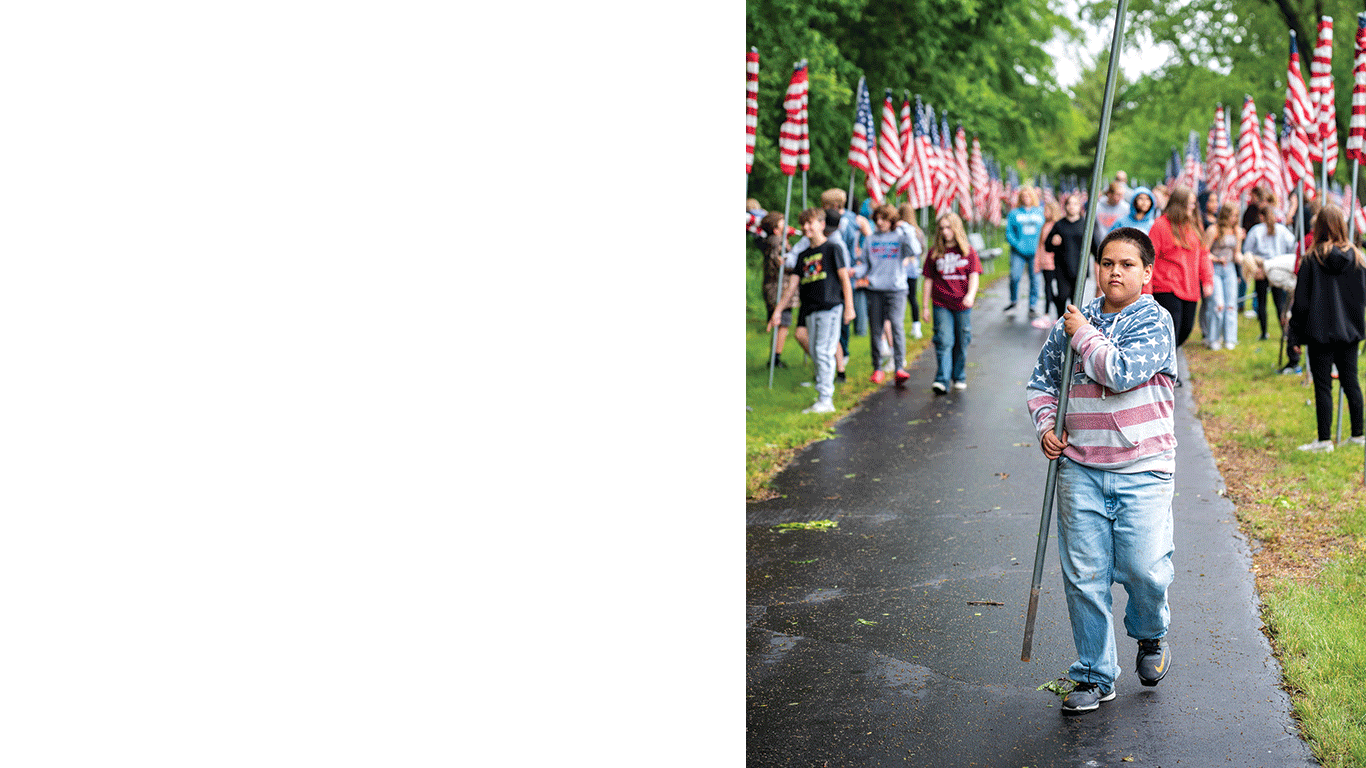 Young person walks along a paved path carrying a flagpole, with many people and American flags lining both sides.