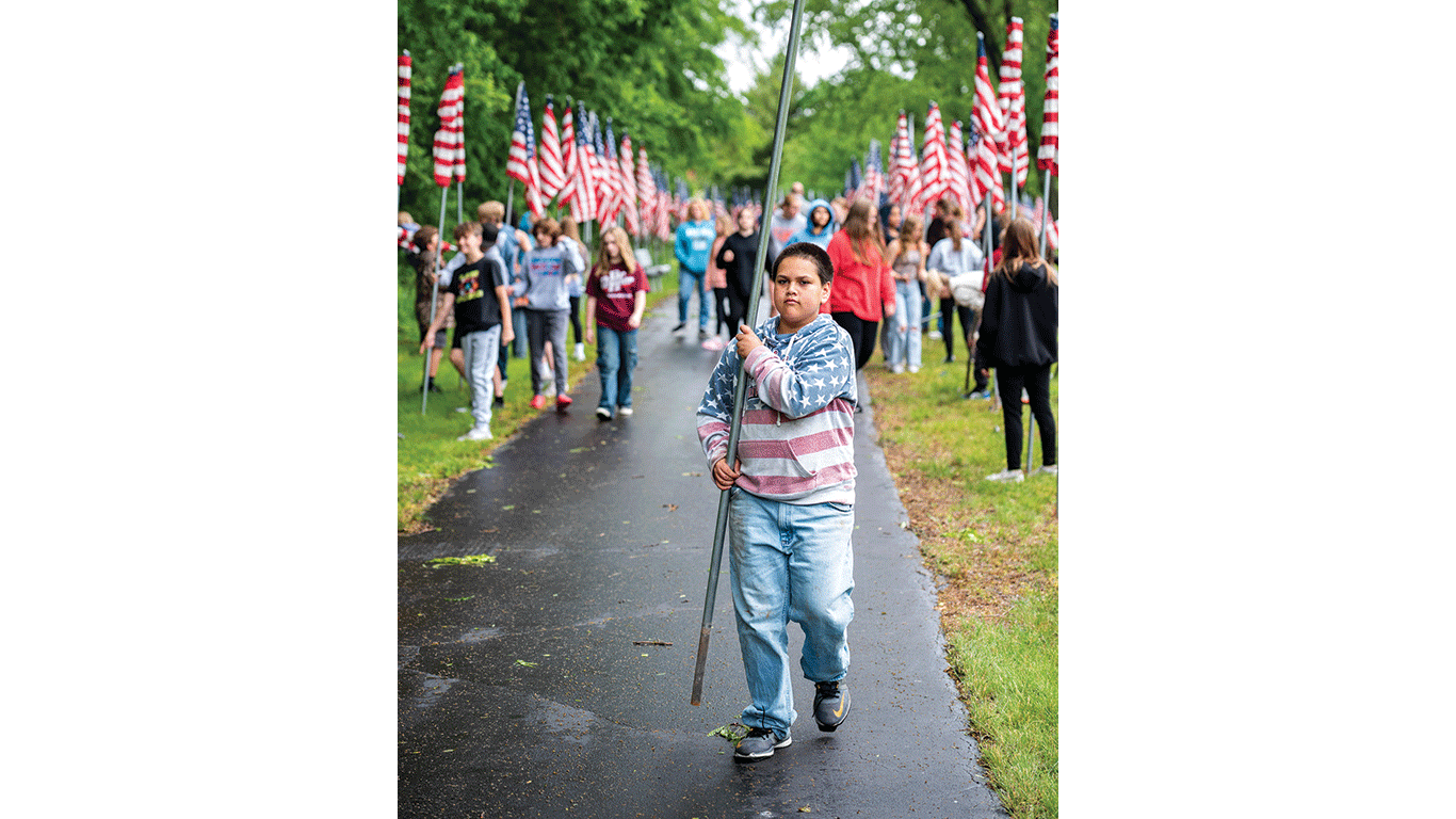 Young person walks along a paved path carrying a flagpole, with many people and American flags lining both sides.