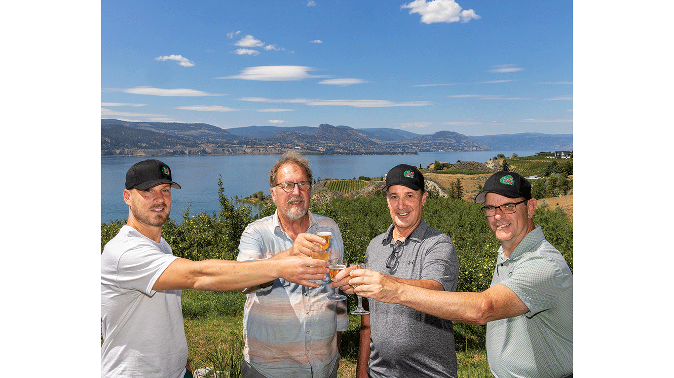 Four people raise glasses together outdoors, with a lake and hills in the background.