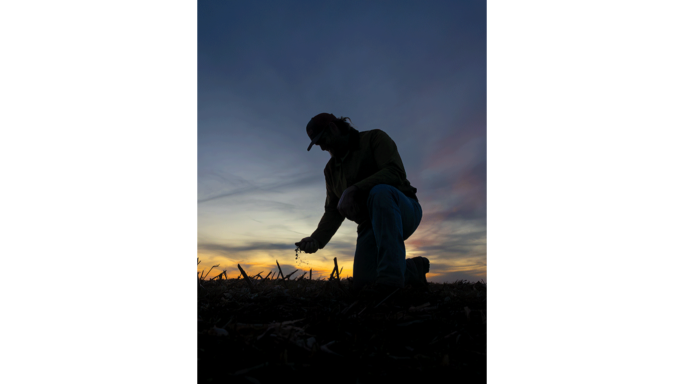Silhouette of a person kneeling in a field, examining soil at sunset under a wide, clouded sky.