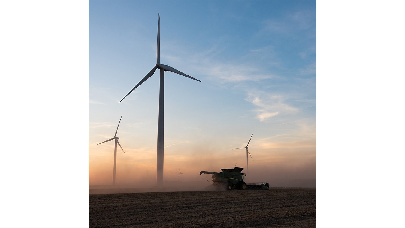 A combine harvests a dusty field at sunset with large wind turbines in the background.