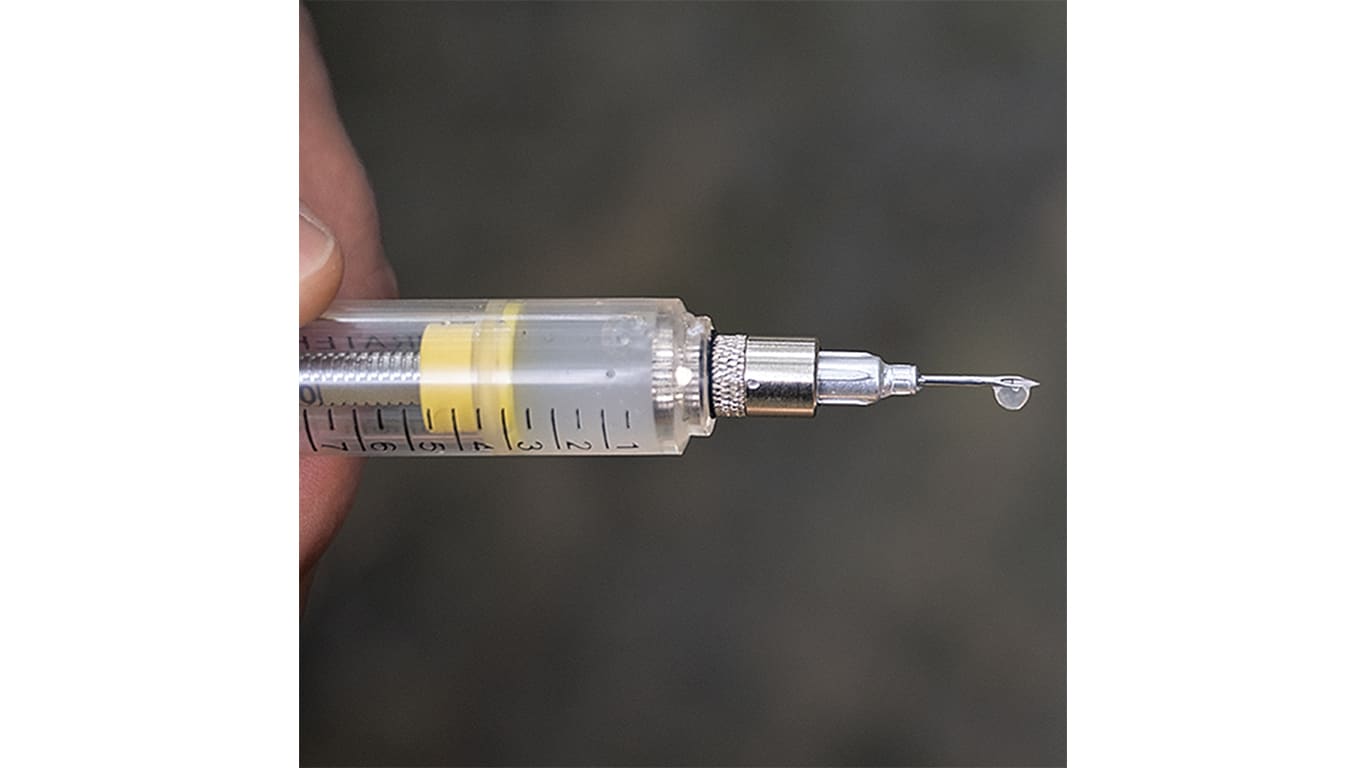 A hand holding a syringe with a metal needle tip and a small droplet forming at the end.
