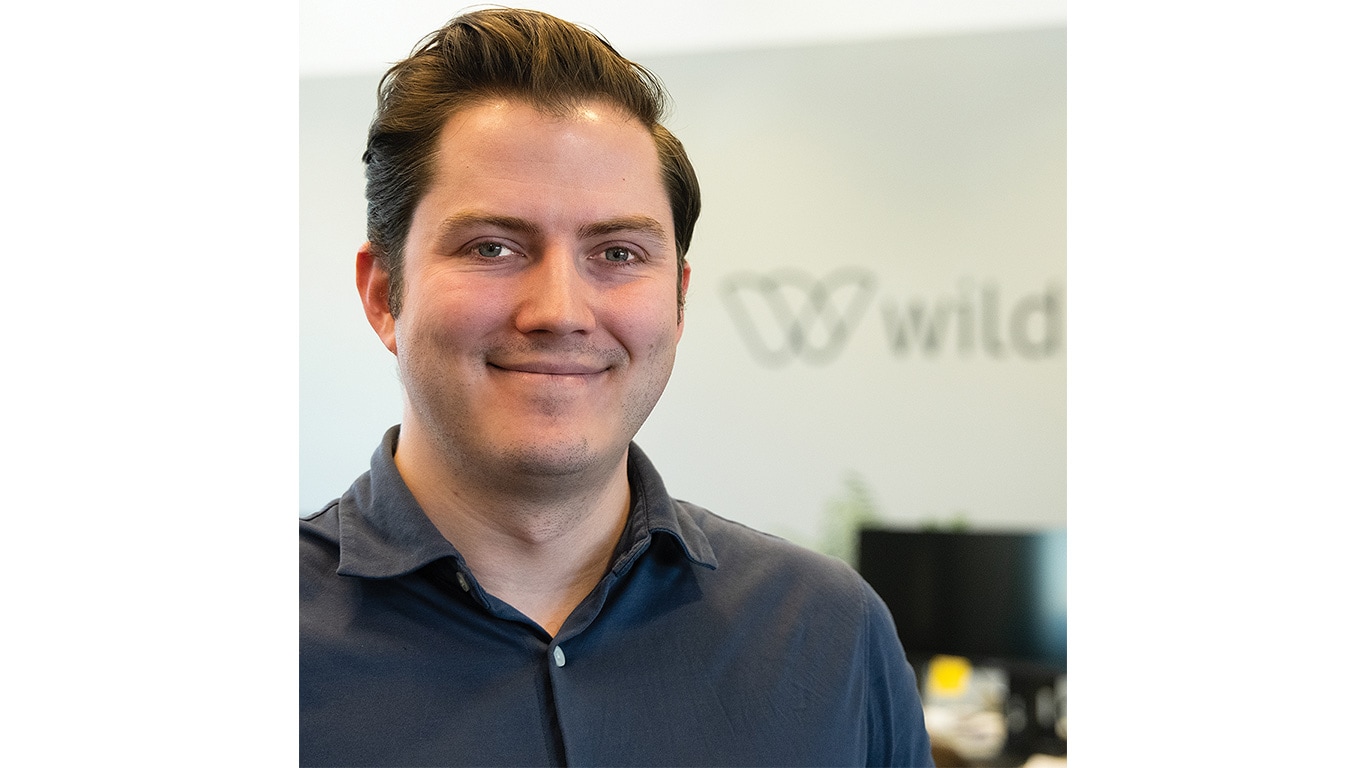 Person in a dark shirt standing in an office with a company logo on the wall.