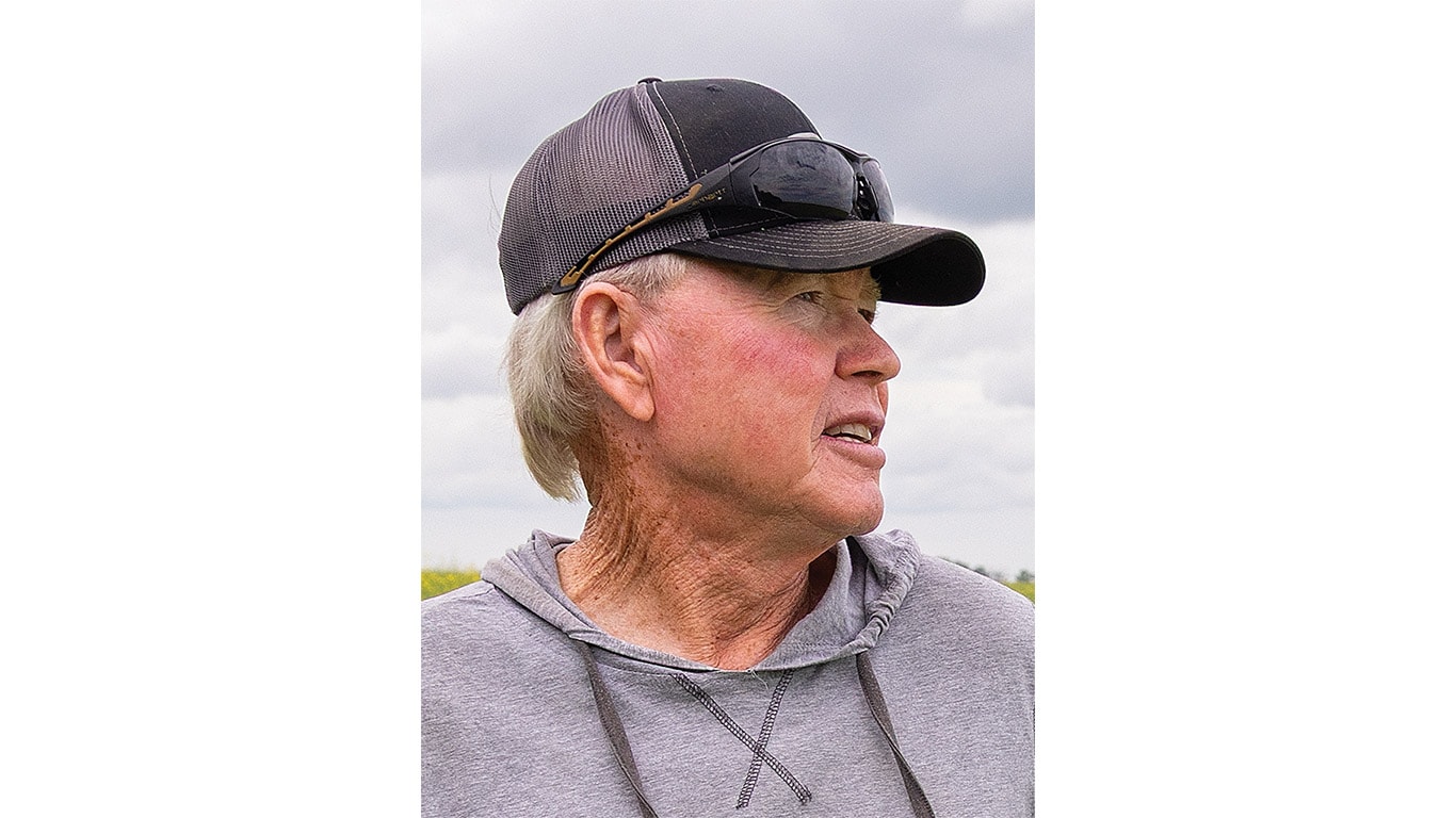A farmer in a baseball cap and hoodie stands in an open field under a cloudy sky.
