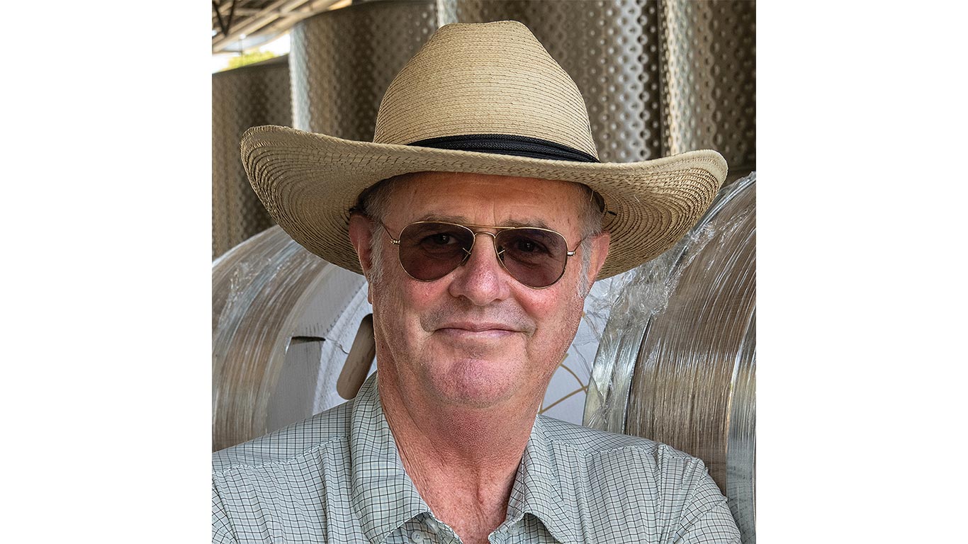 A person wearing a straw hat stands inside a winery with stainless steel fermentation tanks behind them.