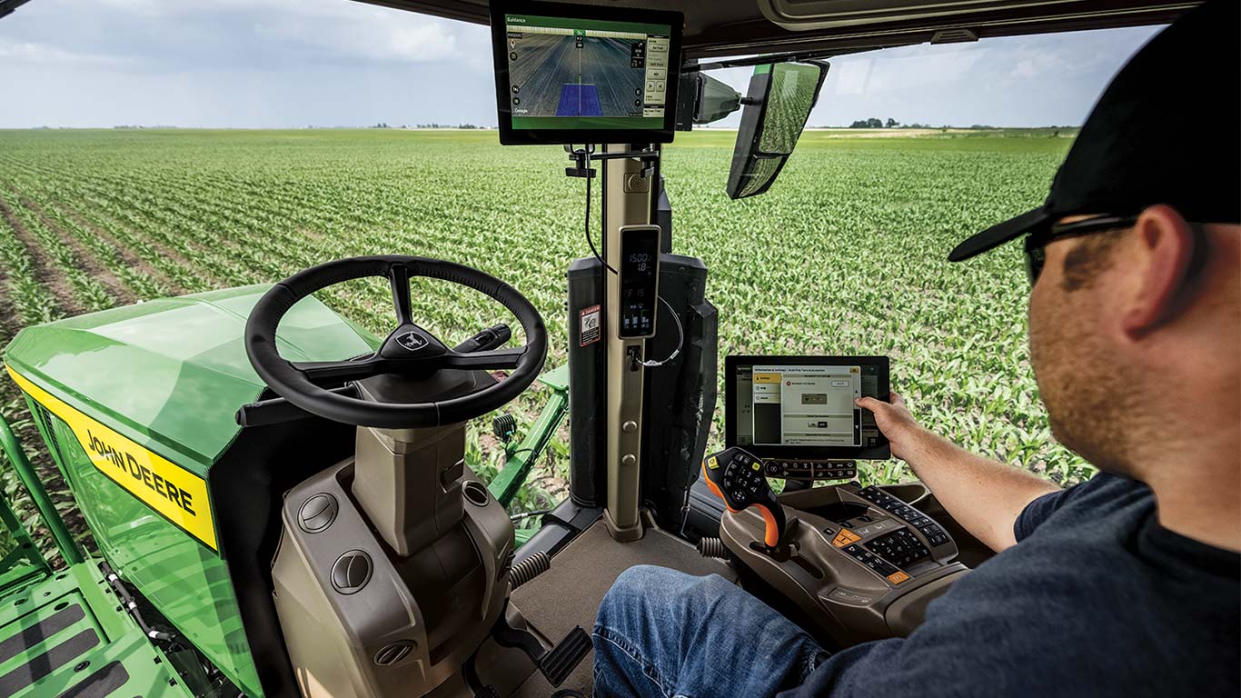 Tractor cab view showing a person using touchscreen controls while overlooking a cornfield.