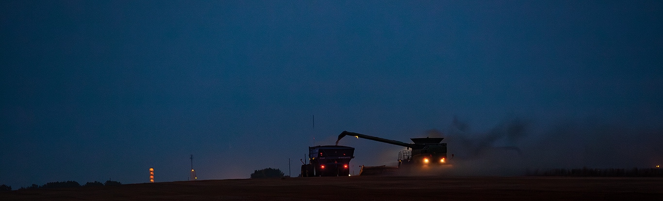 Combine harvester unloading grain into a tractor at dusk with lights glowing.