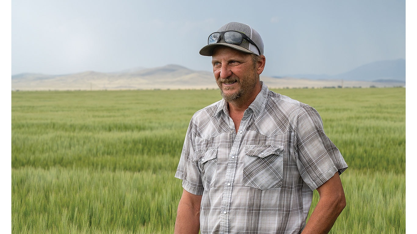 Owner standing in a lush barley field with a scenic mountain range in the background.