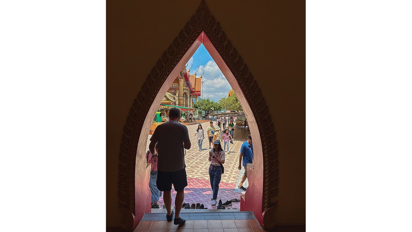 View through an ornate archway to a courtyard with people walking near a temple under blue sky.