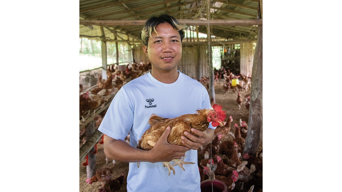 Person in a white shirt holding a brown chicken inside a large poultry house with many hens.