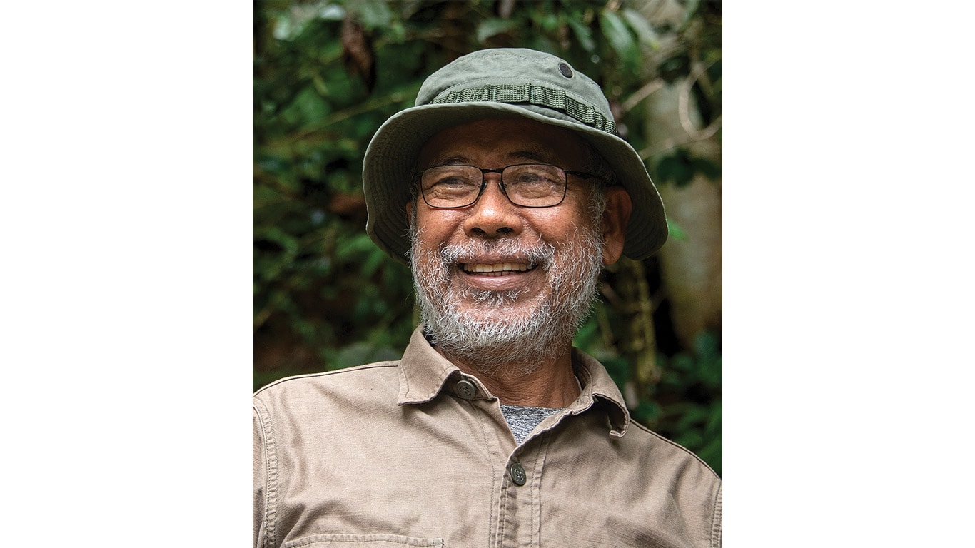 Person wearing a green bucket hat and tan button-up shirt standing outdoors near greenery.
