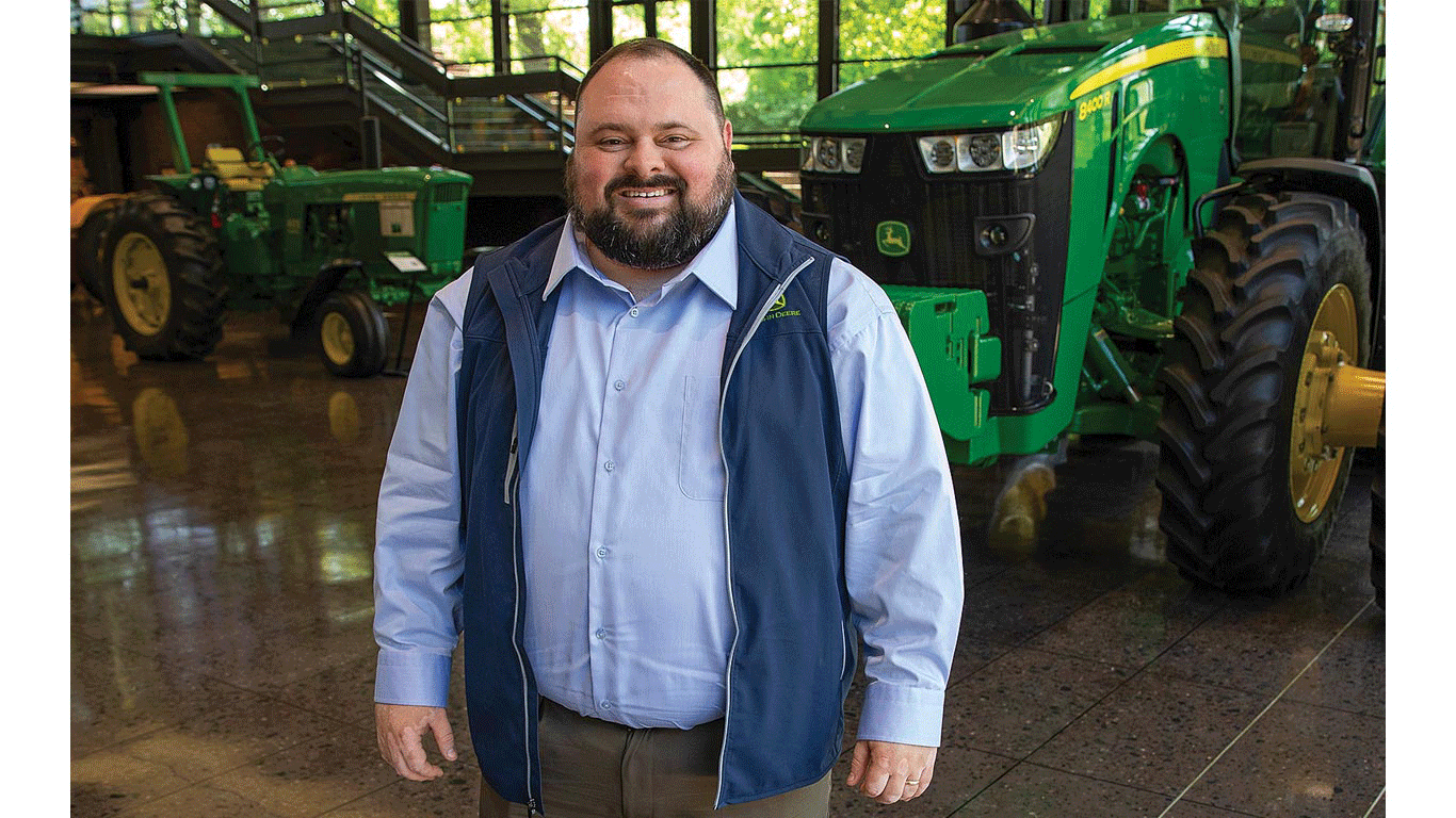 Person in a blue vest standing indoors near green tractors on a polished floor.