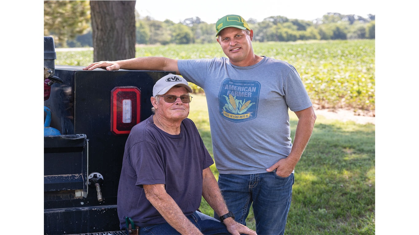 Two people near a black truck with one sitting and one standing in front of a green crop field.