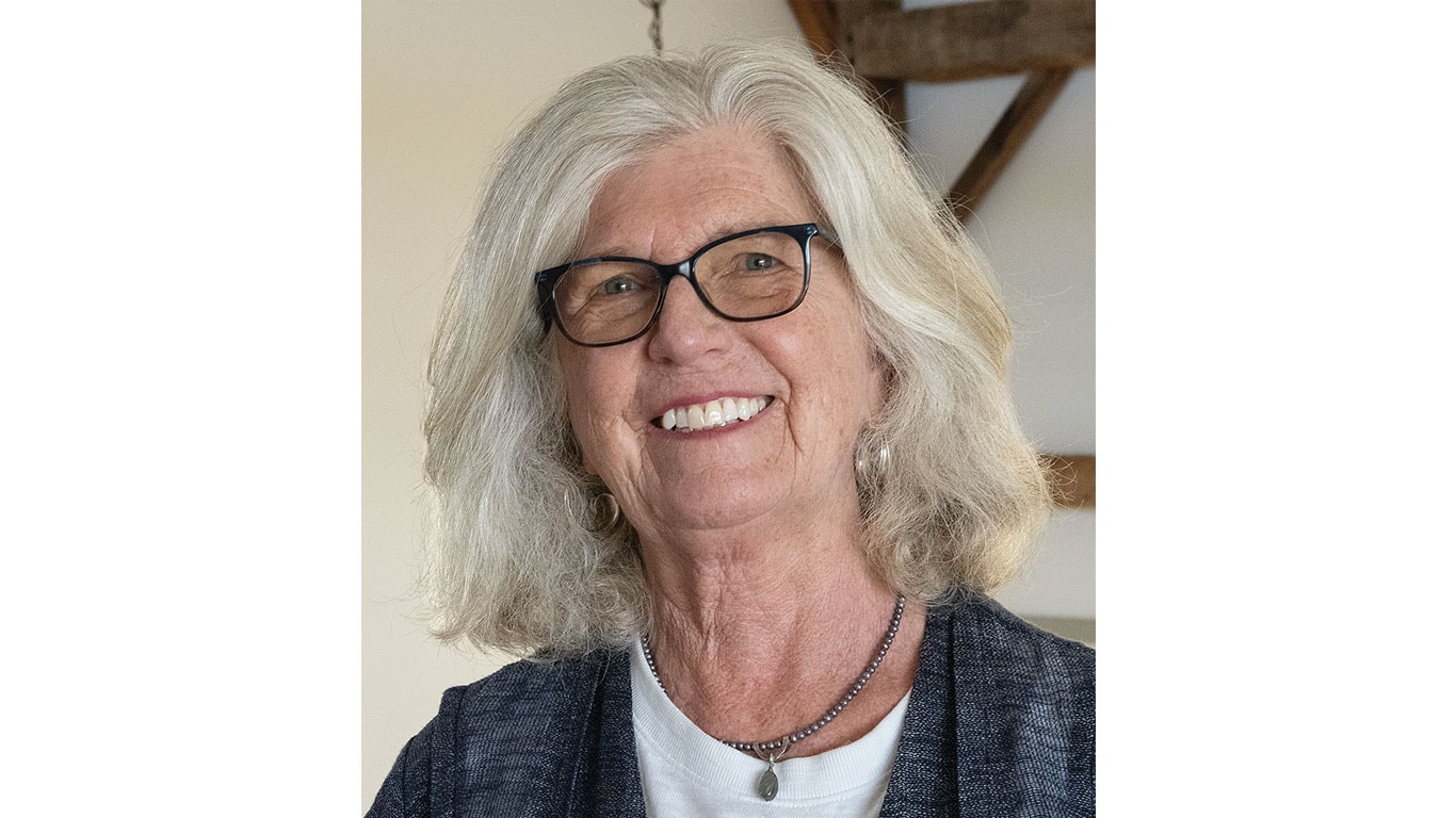 Person with shoulder-length gray hair wearing a dark cardigan and layered necklaces indoors.