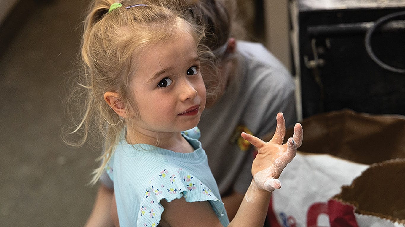 Child with flour-covered hands near an open feed bag indoors.
