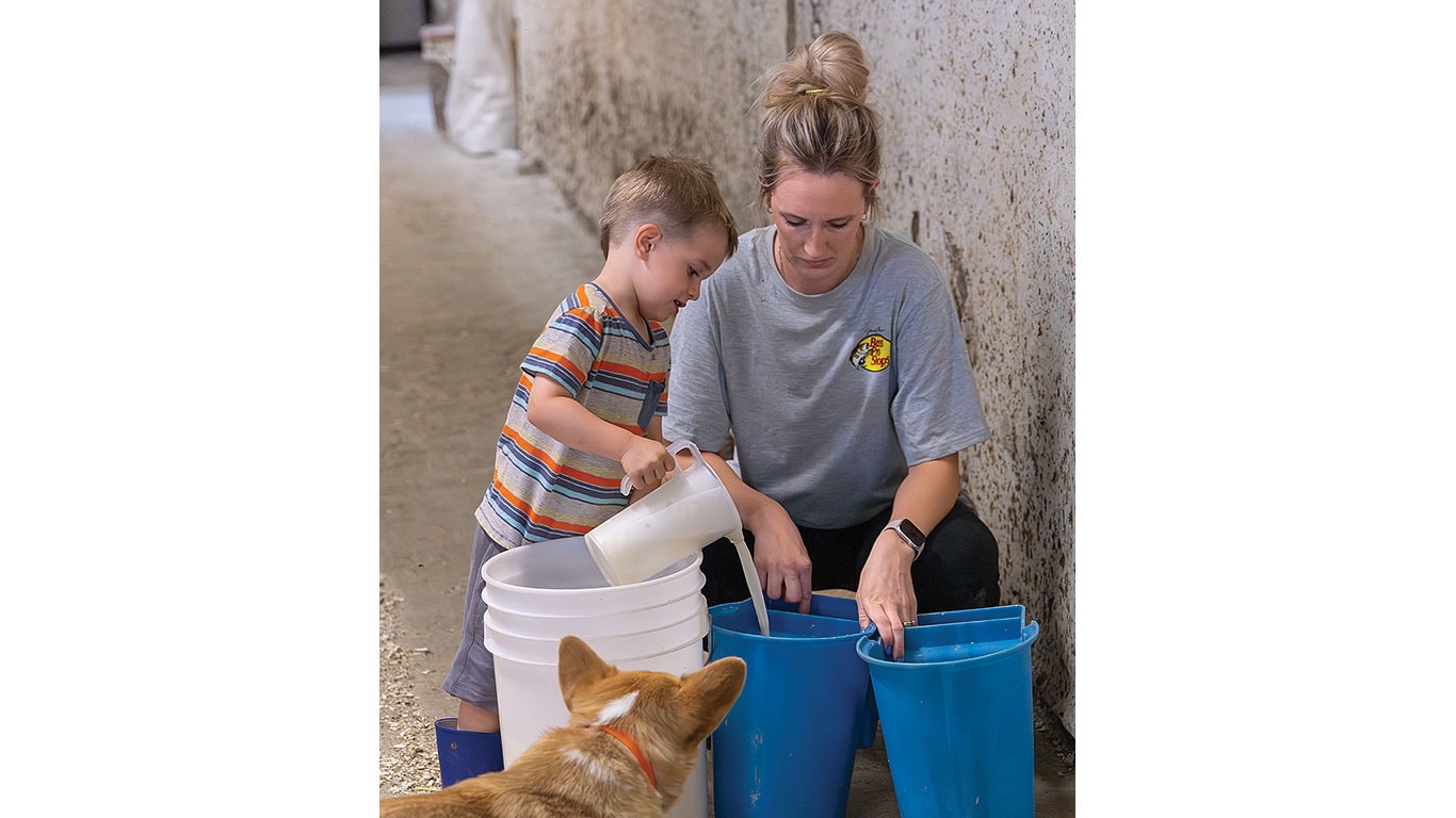 Adult and child filling blue and white buckets with feed as a dog watches nearby.