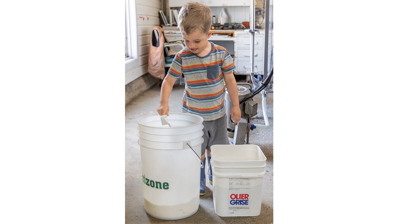 Child standing indoors with two white buckets, one labeled Olier Grise, holding a scoop.